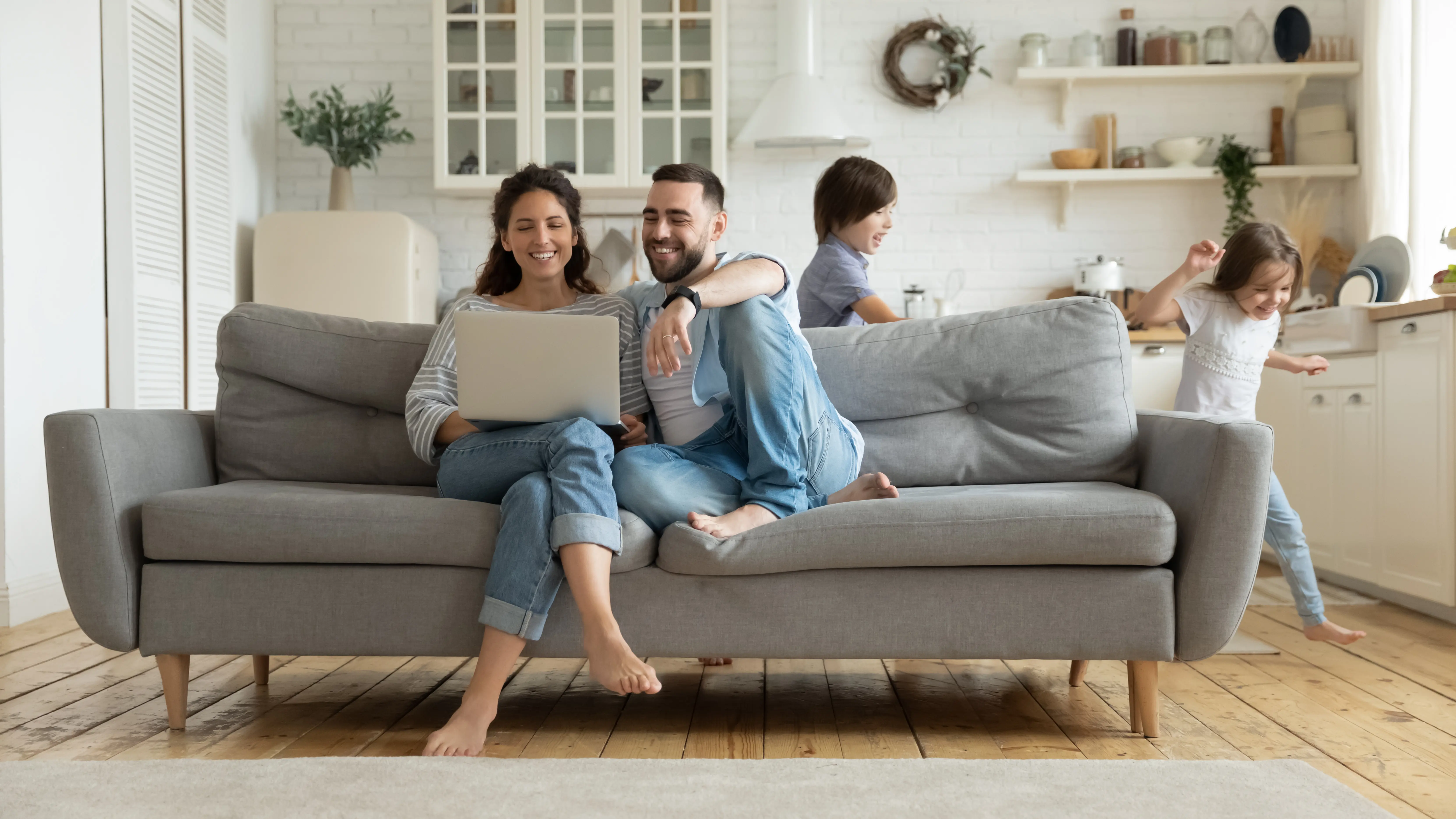 A husband and wife, smiling, sitting on a couch in their home looking at a computer while kids run in the background.